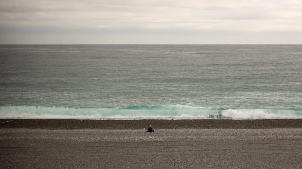 Man sittin on very grey beach alone, with a very grey ocean with very grey waves rolling in towards him.