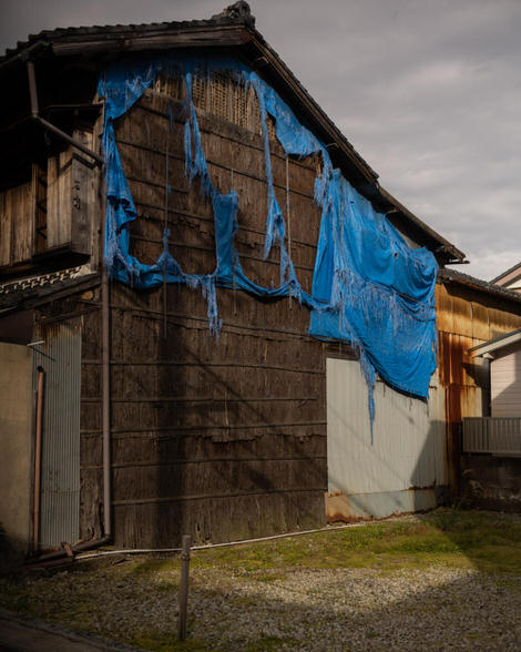 Side of an old Japanese building, tile roof, falling apart, patched up with wind-ripped blue tarp, itself also falling apart - just a mess of a thing in beautiful early mornig light