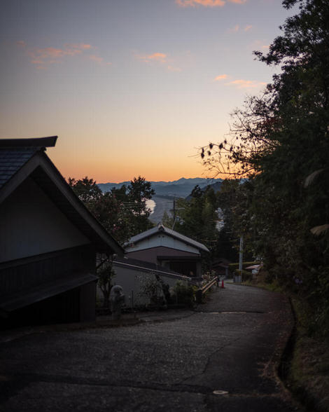 Late-day orange glow towards the horizon, coming down off the final pass, Matsumoto Tōge, Japanese style houses in the foreground, concrete road leading down into town.
