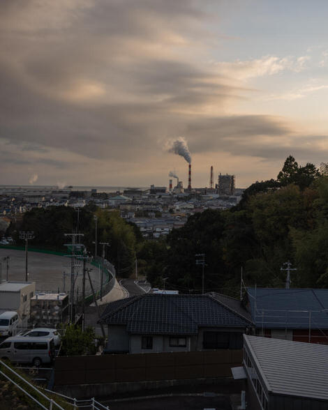 A industrial landscape as seen from a high-ish vantage point; chimney stacks in far distance, billowing smoke against a late-afternoon sky of blue and white and warm colored clouds.