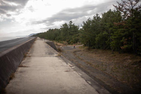 Walking atop a seemingly endless seawall, stretching straight towards the horizon, to the right in the far edge of the frame is a man hitting a tennis ball with a racket, mid-swing.